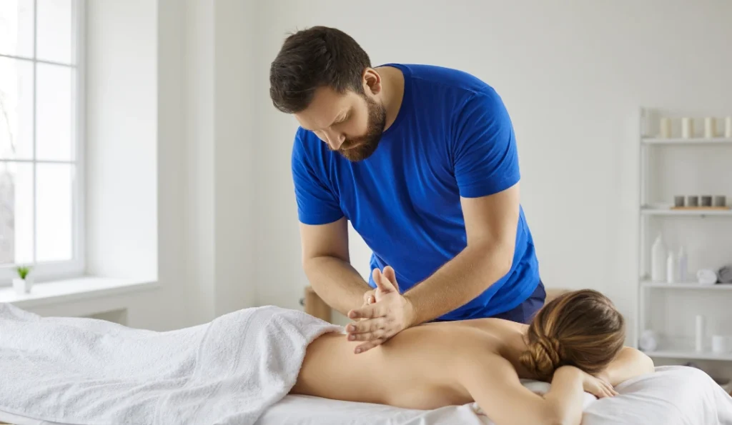 A bearded massage therapist in a blue shirt is shown giving a back massage to a woman lying on her stomach and draped with a white towel. Natural light fills the room from a nearby window. This depicts a massage service, offered at Serenity Head Spas.
