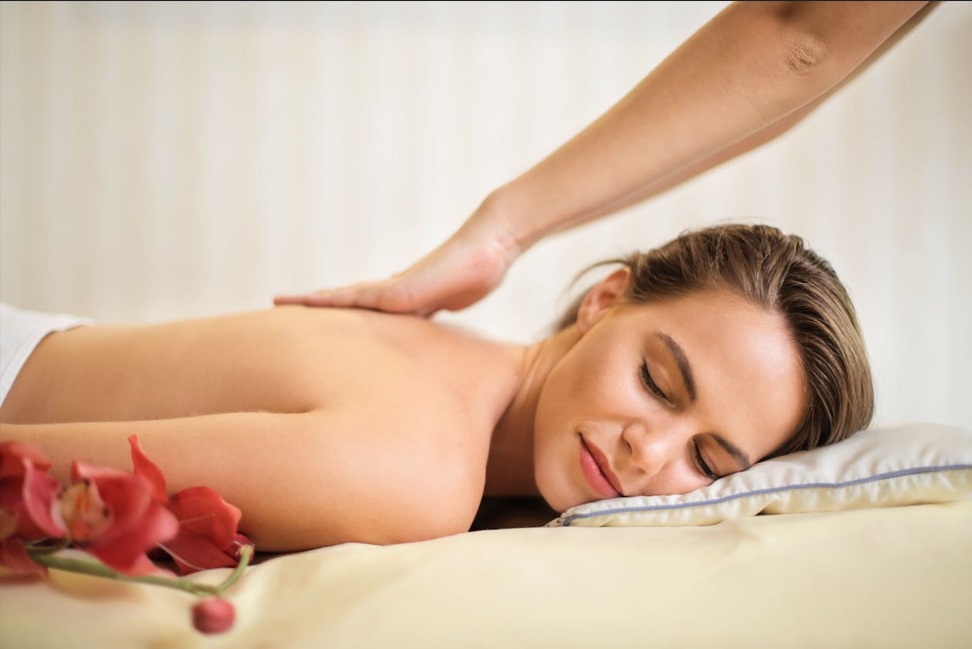 A fair-skinned woman with her eyes closed rests on her stomach on a massage table, a white towel draped around her. A massage therapist's arm and hand are gently placed on her upper back. A vibrant red orchid lies in the foreground. This image represent a relaxing massage offered at Serenity Head Spas.