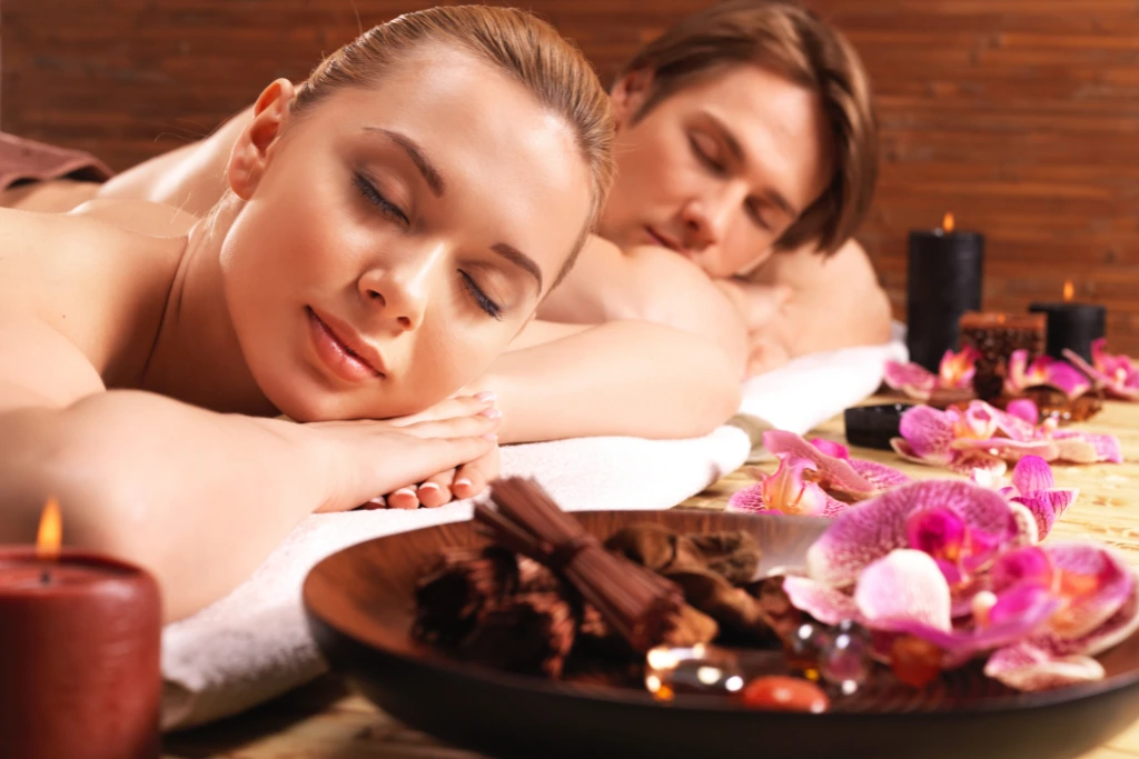 A serene spa scene shows a fair-skinned couple with their eyes closed, seemingly enjoying a relaxing experience at Serenity Head Spas. The foreground features a wooden bowl with aromatic elements and vibrant pink orchids, while soft candlelight adds to the tranquil atmosphere. This could represent a couples massage offered by Serenity Head Spas.