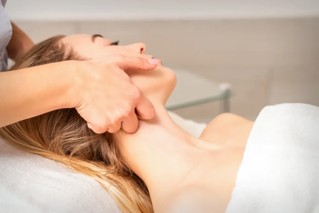 A woman receiving a manual lymphatic drainage massage on her face and neck. The massage therapist's hands are visible applying gentle pressure.